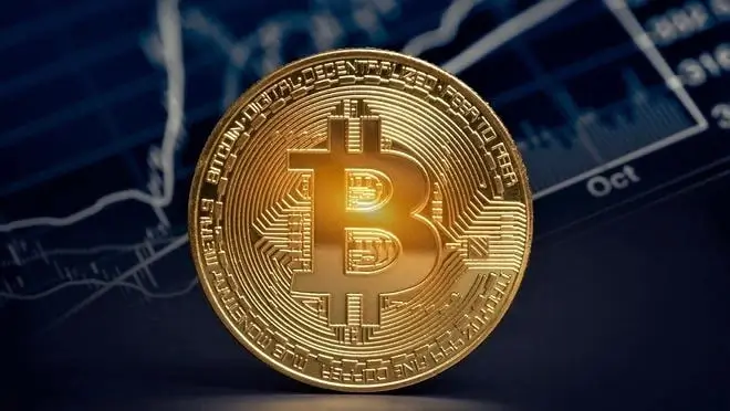 A view of a gold Bitcoin coin against a blue background with charts